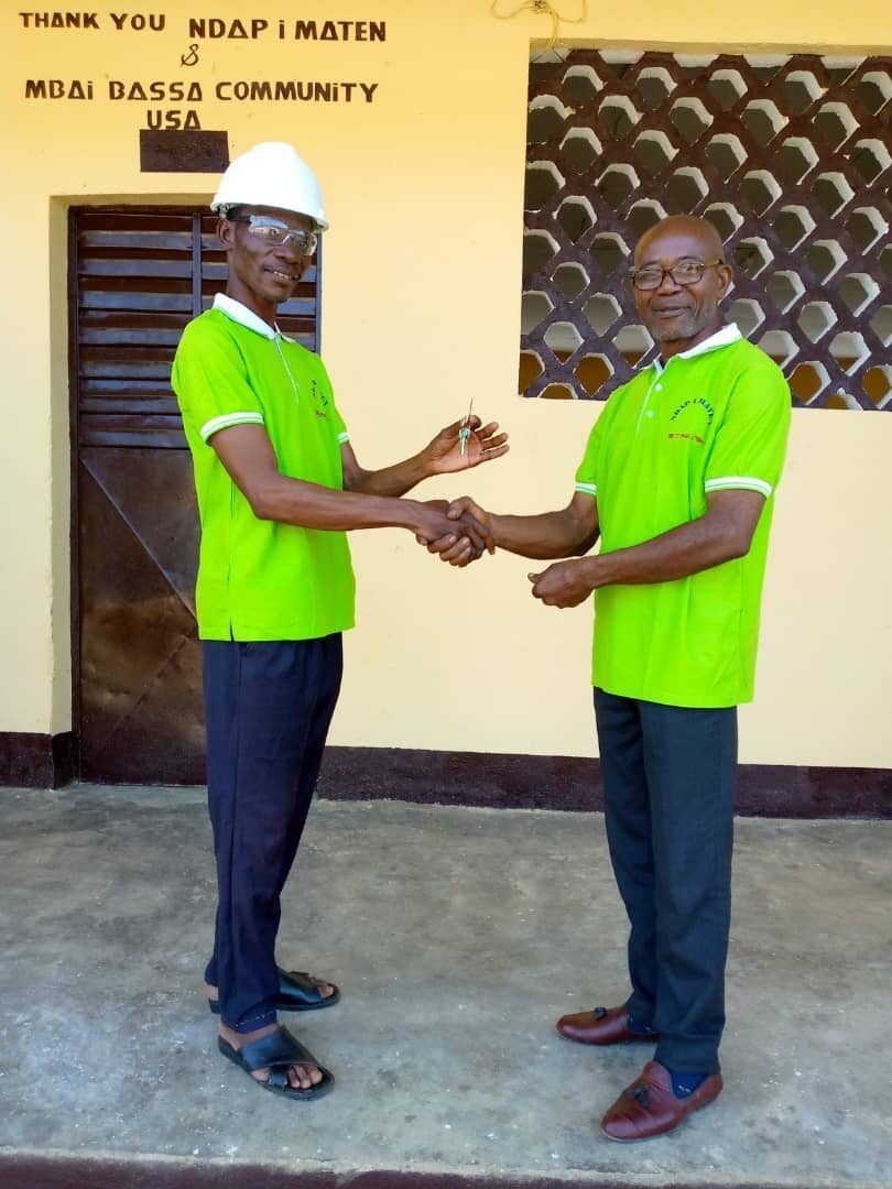 Two men in Ndap I Maten shirts shake hands in front of a thank-you sign at the renovated Ngonga school